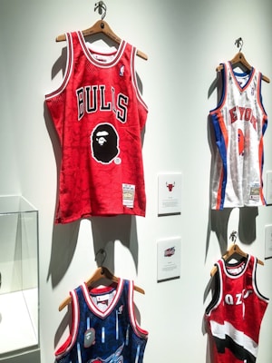 A display of several basketball jerseys hanging on a wall, each on wooden hangers. The prominent jersey is red with the word 'BULLS' and a graphic face logo. Other jerseys include a white New York jersey with blue and red accents and a blue jersey with intricate patterns.