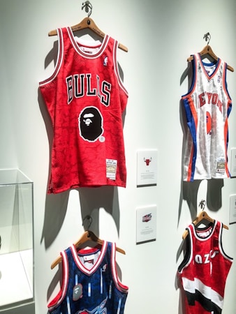 A display of several basketball jerseys hanging on a wall, each on wooden hangers. The prominent jersey is red with the word 'BULLS' and a graphic face logo. Other jerseys include a white New York jersey with blue and red accents and a blue jersey with intricate patterns.