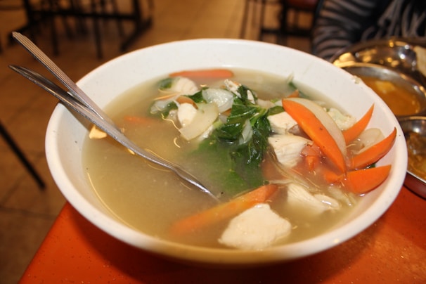 A bowl of clear broth soup containing sliced carrots, leafy green vegetables, and chunks of white meat, likely chicken. Two silver spoons are placed in the bowl. The bowl is on an orange table, and there are stainless steel dishes in the background.