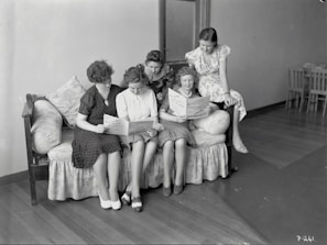 Five women are gathered together on a sofa, intently reading papers or pamphlets. The scene appears to be from a past era, as suggested by the vintage style of their clothing. The room is simple with wooden flooring and minimal furnishings, including a closed door and a few chairs in the background.