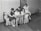 Five women are gathered together on a sofa, intently reading papers or pamphlets. The scene appears to be from a past era, as suggested by the vintage style of their clothing. The room is simple with wooden flooring and minimal furnishings, including a closed door and a few chairs in the background.