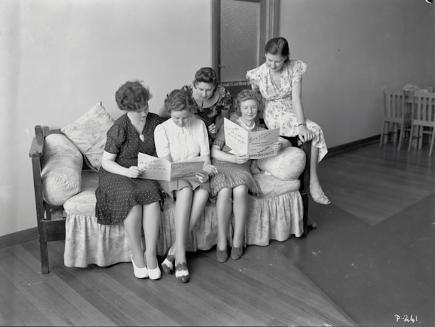 Five women are gathered together on a sofa, intently reading papers or pamphlets. The scene appears to be from a past era, as suggested by the vintage style of their clothing. The room is simple with wooden flooring and minimal furnishings, including a closed door and a few chairs in the background.