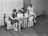 Five women are gathered together on a sofa, intently reading papers or pamphlets. The scene appears to be from a past era, as suggested by the vintage style of their clothing. The room is simple with wooden flooring and minimal furnishings, including a closed door and a few chairs in the background.