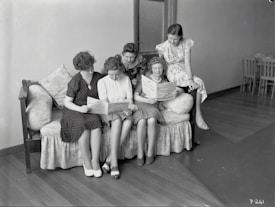 Five women are gathered together on a sofa, intently reading papers or pamphlets. The scene appears to be from a past era, as suggested by the vintage style of their clothing. The room is simple with wooden flooring and minimal furnishings, including a closed door and a few chairs in the background.