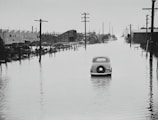 A vintage car is partially submerged on a flooded street, flanked by utility poles and industrial buildings. The area appears to be heavily inundated by water with reflections of structures in the floodwater. The sky is overcast, adding a somber feel to the scene.