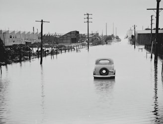A vintage car is partially submerged on a flooded street, flanked by utility poles and industrial buildings. The area appears to be heavily inundated by water with reflections of structures in the floodwater. The sky is overcast, adding a somber feel to the scene.