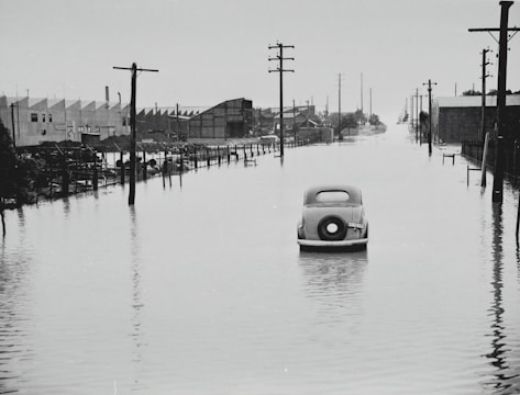 A vintage car is partially submerged on a flooded street, flanked by utility poles and industrial buildings. The area appears to be heavily inundated by water with reflections of structures in the floodwater. The sky is overcast, adding a somber feel to the scene.