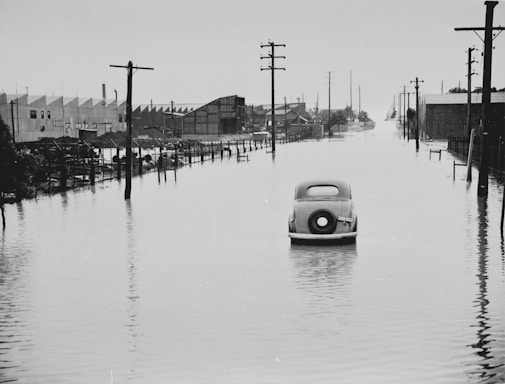 A vintage car is partially submerged on a flooded street, flanked by utility poles and industrial buildings. The area appears to be heavily inundated by water with reflections of structures in the floodwater. The sky is overcast, adding a somber feel to the scene.
