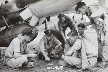 A group of six men in military uniforms are sitting and crouching under an aircraft, likely on a military base. They are engaged in a card game, with some holding cards and others focused on the game. The setting suggests a moment of leisure amidst a military environment, possibly during World War II.