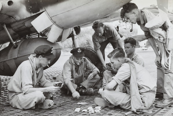 A group of six men in military uniforms are sitting and crouching under an aircraft, likely on a military base. They are engaged in a card game, with some holding cards and others focused on the game. The setting suggests a moment of leisure amidst a military environment, possibly during World War II.