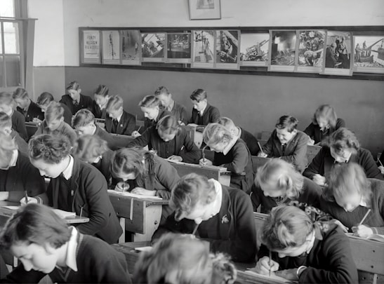 A classroom filled with students deeply focused on writing in their notebooks. The students, dressed uniformly, are seated in wooden desks lined in neat rows. The room is adorned with educational posters and has a large window allowing natural light to illuminate the space.