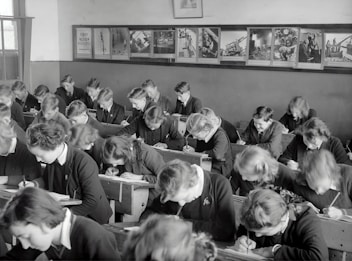A classroom filled with students deeply focused on writing in their notebooks. The students, dressed uniformly, are seated in wooden desks lined in neat rows. The room is adorned with educational posters and has a large window allowing natural light to illuminate the space.