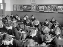 A classroom filled with students deeply focused on writing in their notebooks. The students, dressed uniformly, are seated in wooden desks lined in neat rows. The room is adorned with educational posters and has a large window allowing natural light to illuminate the space.