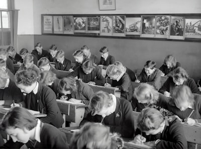 A classroom filled with students deeply focused on writing in their notebooks. The students, dressed uniformly, are seated in wooden desks lined in neat rows. The room is adorned with educational posters and has a large window allowing natural light to illuminate the space.