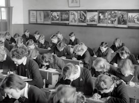 A classroom filled with students deeply focused on writing in their notebooks. The students, dressed uniformly, are seated in wooden desks lined in neat rows. The room is adorned with educational posters and has a large window allowing natural light to illuminate the space.