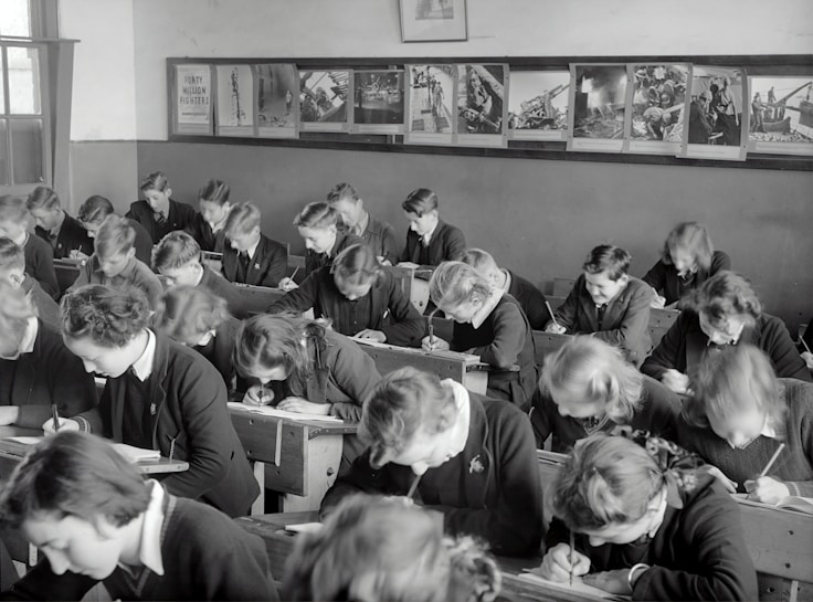 A classroom filled with students deeply focused on writing in their notebooks. The students, dressed uniformly, are seated in wooden desks lined in neat rows. The room is adorned with educational posters and has a large window allowing natural light to illuminate the space.
