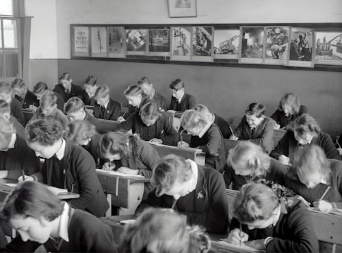 A classroom filled with students deeply focused on writing in their notebooks. The students, dressed uniformly, are seated in wooden desks lined in neat rows. The room is adorned with educational posters and has a large window allowing natural light to illuminate the space.
