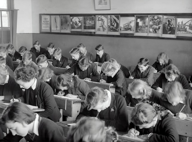 A classroom filled with students deeply focused on writing in their notebooks. The students, dressed uniformly, are seated in wooden desks lined in neat rows. The room is adorned with educational posters and has a large window allowing natural light to illuminate the space.