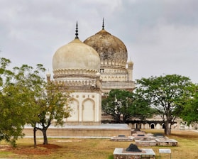 A historic mausoleum with intricate architectural design and large domes stands surrounded by trees. The structure exhibits ornate decoration with a blend of white and weathered stone. Below the mausoleum, several gravestones are seen on a grass-covered ground.