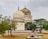 A historic mausoleum with intricate architectural design and large domes stands surrounded by trees. The structure exhibits ornate decoration with a blend of white and weathered stone. Below the mausoleum, several gravestones are seen on a grass-covered ground.