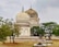 A historic mausoleum with intricate architectural design and large domes stands surrounded by trees. The structure exhibits ornate decoration with a blend of white and weathered stone. Below the mausoleum, several gravestones are seen on a grass-covered ground.
