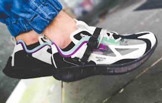 A pair of stylish Reebok sneakers worn by someone in blue jeans. The sneakers feature a combination of colors including white, black, purple, and mint green with a visible logo on the side. They have a modern design with a sleek, sporty aesthetic.