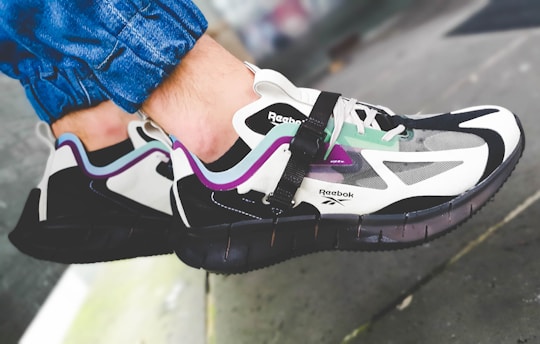 A pair of stylish Reebok sneakers worn by someone in blue jeans. The sneakers feature a combination of colors including white, black, purple, and mint green with a visible logo on the side. They have a modern design with a sleek, sporty aesthetic.
