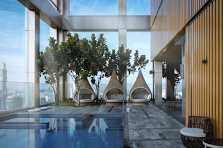 A modern outdoor space features three wicker teardrop-shaped lounge chairs with cushions, positioned in front of tall green plants. The area includes a pool with clear blue water reflecting the sky. The architecture includes glass walls and a building with a golden-hued metallic exterior, creating a blend of natural and urban elements.