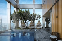 A modern outdoor space features three wicker teardrop-shaped lounge chairs with cushions, positioned in front of tall green plants. The area includes a pool with clear blue water reflecting the sky. The architecture includes glass walls and a building with a golden-hued metallic exterior, creating a blend of natural and urban elements.