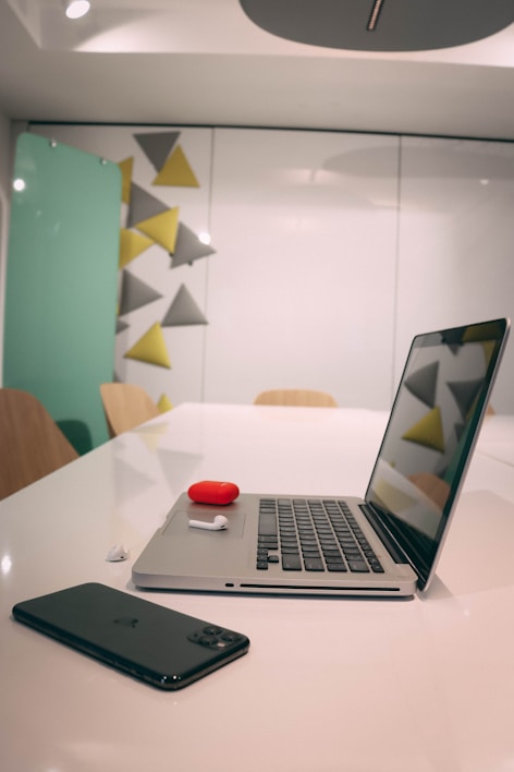 A modern, minimalistic office setting with a laptop, smartphone, and wireless earbuds displayed on a polished white table. The backdrop features a geometric decoration of gray and yellow triangular shapes. The atmosphere is tidy and contemporary.