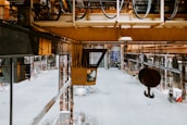 An industrial warehouse with high ceilings, metal beams, and various machinery. There are large hooks suspended from the ceiling, and the space is open with some equipment and cables visible. The floor is mostly empty, creating a spacious feel.