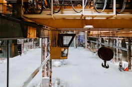 An industrial warehouse with high ceilings, metal beams, and various machinery. There are large hooks suspended from the ceiling, and the space is open with some equipment and cables visible. The floor is mostly empty, creating a spacious feel.