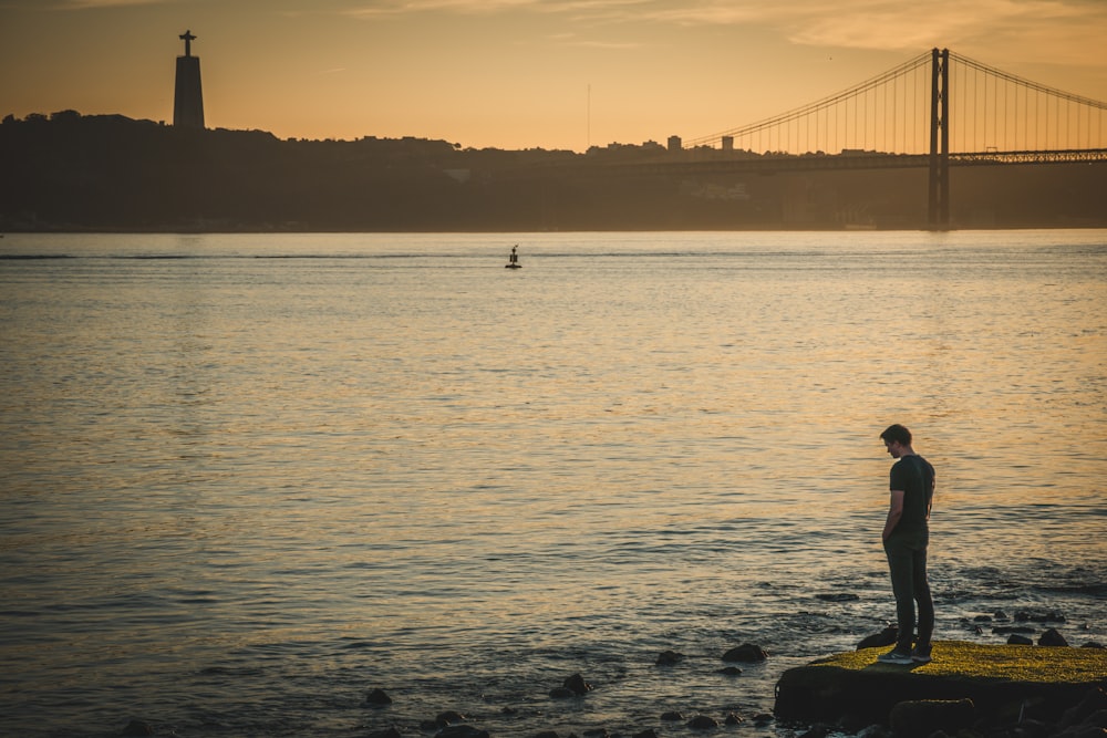 man in white shirt sitting on rock in front of sea during sunset