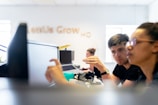Two people are engaged in a discussion while working at a desk with computers in an open office setting. A blurred background contains other people working and a sign on the wall with text partially visible.