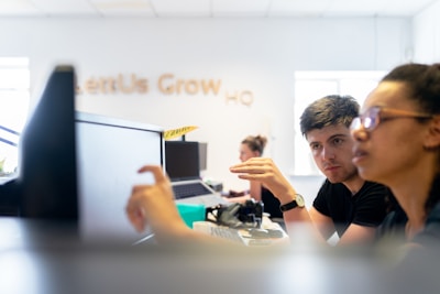 Two people are engaged in a discussion while working at a desk with computers in an open office setting. A blurred background contains other people working and a sign on the wall with text partially visible.