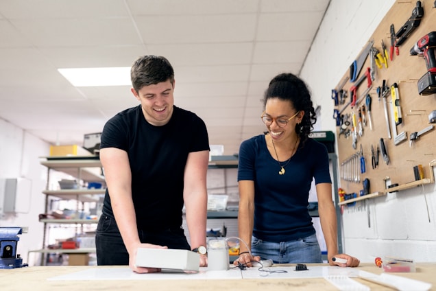 Two people are engaged in a collaborative workshop setting. They are smiling and appear to be working on a project with various tools and equipment on the table. The walls feature an organized display of tools.