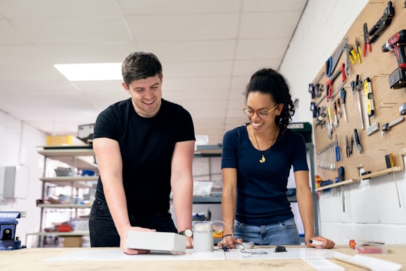 Two people are engaged in a collaborative workshop setting. They are smiling and appear to be working on a project with various tools and equipment on the table. The walls feature an organized display of tools.