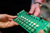 Two hands are holding a green electronic circuit board. The board contains various components such as capacitors, resistors, and connectors, with a label that reads 'LettUS Grow'. It appears to be part of an electronic device or system.