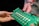 Two hands are holding a green electronic circuit board. The board contains various components such as capacitors, resistors, and connectors, with a label that reads 'LettUS Grow'. It appears to be part of an electronic device or system.