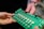 Two hands are holding a green electronic circuit board. The board contains various components such as capacitors, resistors, and connectors, with a label that reads 'LettUS Grow'. It appears to be part of an electronic device or system.