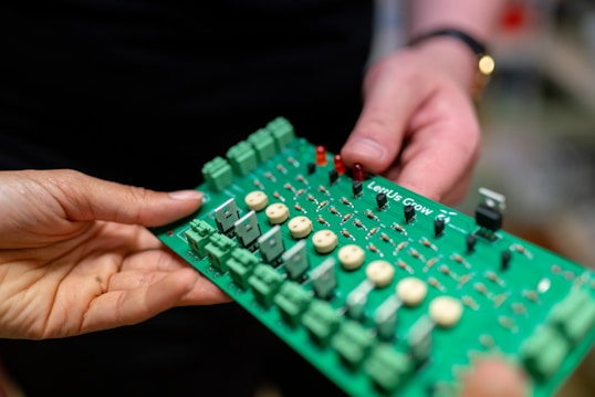 Two hands are holding a green electronic circuit board. The board contains various components such as capacitors, resistors, and connectors, with a label that reads 'LettUS Grow'. It appears to be part of an electronic device or system.