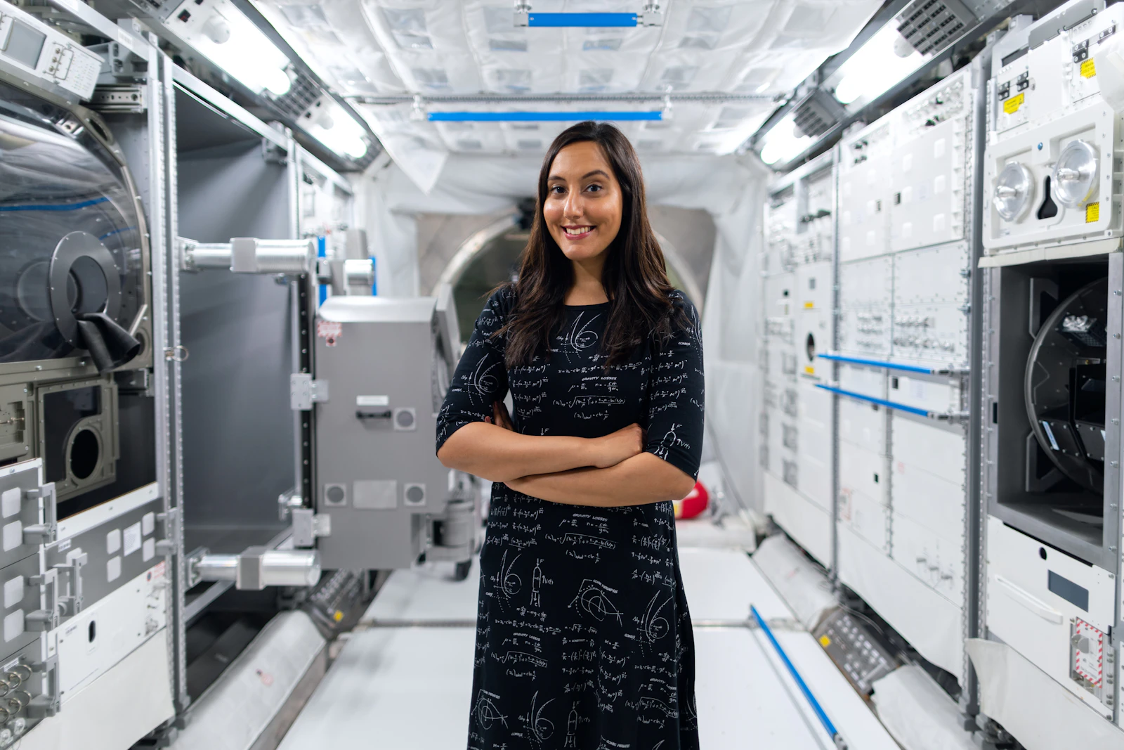 A woman with long dark hair stands smiling with arms crossed inside a space station module mockup, wearing a black dress printed with physics equations.