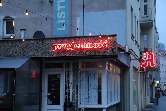 A small urban cafe with a neon sign displaying the word 'przyjemność' in red against a textured facade. There is another red neon letter 'A' on the side of the building. String lights are hung above the entrance, and the area appears to be in a city alley with adjoining buildings.