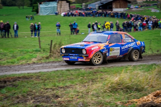 A classic blue and orange rally car is speeding through a muddy track. The vehicle is covered in sponsor decals and is navigating a turn with visible mud splatter on its sides. In the background, a crowd of spectators stands along the edge of the track, observing the race. The setting is a rural area with grassy fields and rustic barns.