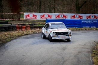 A vintage rally car is taking a sharp turn on a racetrack surrounded by trees. The car is decorated with various sponsor logos, including Castrol, and has a white body with colorful stripes. In the background, there are tire barriers and signs advertising 'Champion's' and an amusement park.