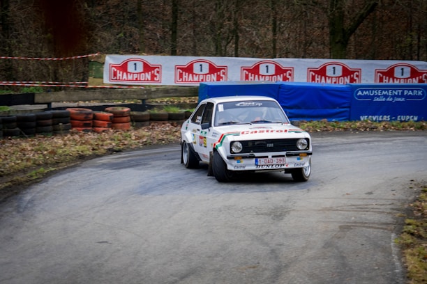 A vintage rally car is taking a sharp turn on a racetrack surrounded by trees. The car is decorated with various sponsor logos, including Castrol, and has a white body with colorful stripes. In the background, there are tire barriers and signs advertising 'Champion's' and an amusement park.