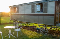 An antique wooden caravan with detailed carvings stands on a grassy field during sunset. The caravan has two shuttered windows and intricate detailing along its side. A white wrought iron table with two matching chairs is situated in the foreground, casting long shadows from the warm evening sun.