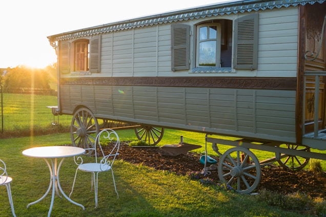 An antique wooden caravan with detailed carvings stands on a grassy field during sunset. The caravan has two shuttered windows and intricate detailing along its side. A white wrought iron table with two matching chairs is situated in the foreground, casting long shadows from the warm evening sun.