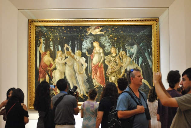 A group of people are observing a large, framed artwork that features several elegantly dressed figures in a classical setting. The painting is mounted on a gallery wall with a simple border and is crowded with visitors taking photos and examining the art.