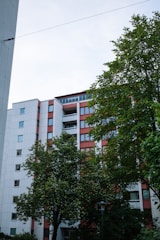 A multistory residential building with a modern design surrounded by green trees. The building has a white and red color scheme with multiple windows and balconies. The sky is slightly overcast, adding to the serene atmosphere.
